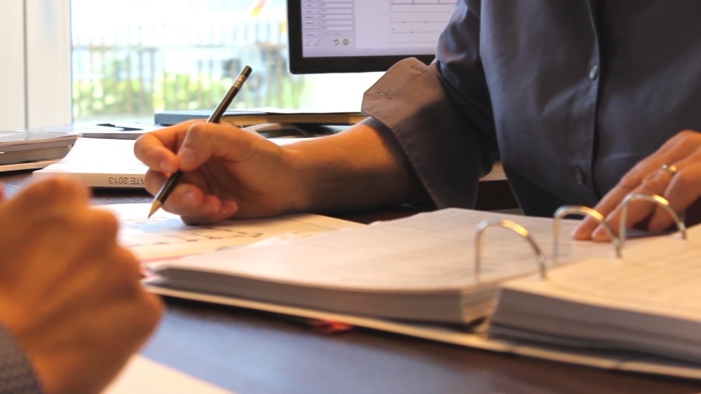 Zwei Personen sitzen sich am Schreibtisch gegenüber, der eine mit einem Ordner. 